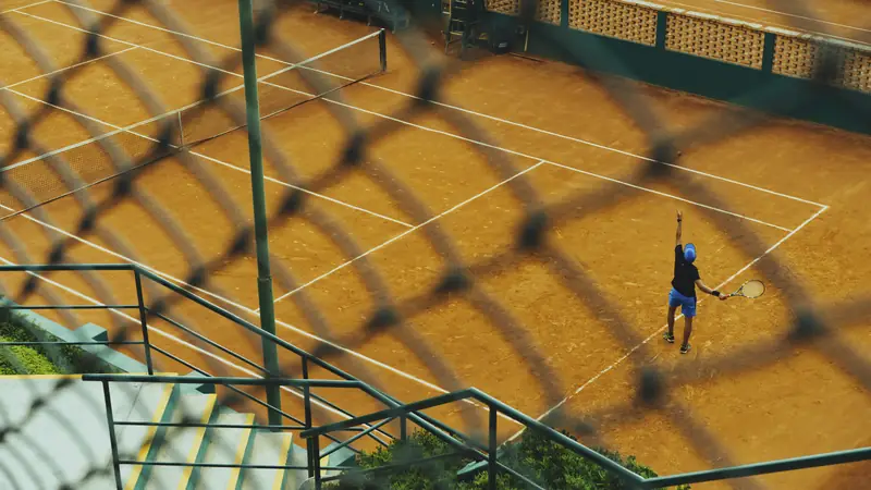 Tennis player serving on a clay court, seen through a wire fence