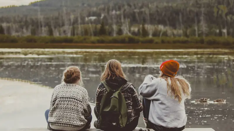 Three friends with backpacks sitting together by a lake